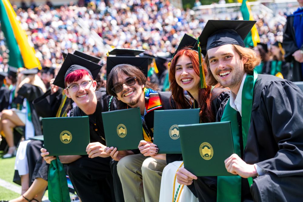 A photo of four graduates sitting down holding up their diploma covers on Commencement.