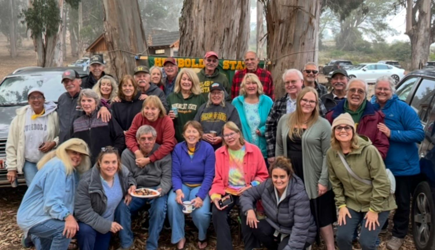 A group of people smiling and posing together in front of trees and parked cars