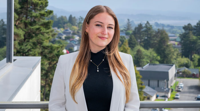 Harmony Switzer-Tryon in a white blazer stands confidently on a balcony, with a view of trees and other campus buildings