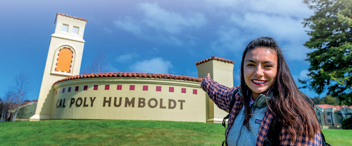 Student wearing red lipstick pointing at the Cal Poly entrance sign