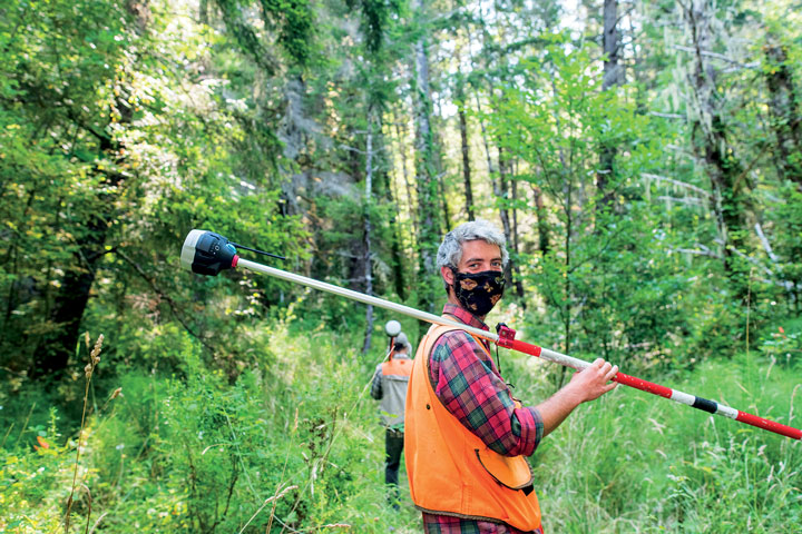 student holding a long peice of equipment in the forest, smiling for the camera