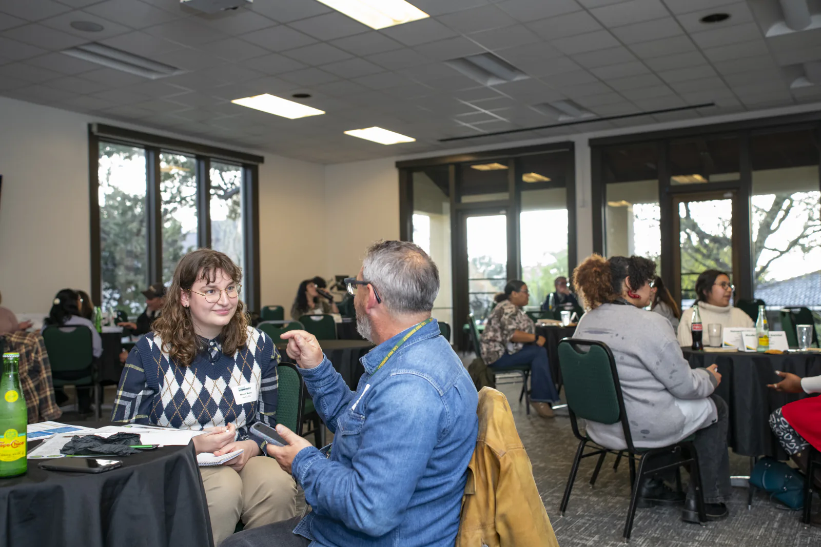 A student listens as an alum gestures during a conversation, with other groups meeting around the Goodwin Forum.