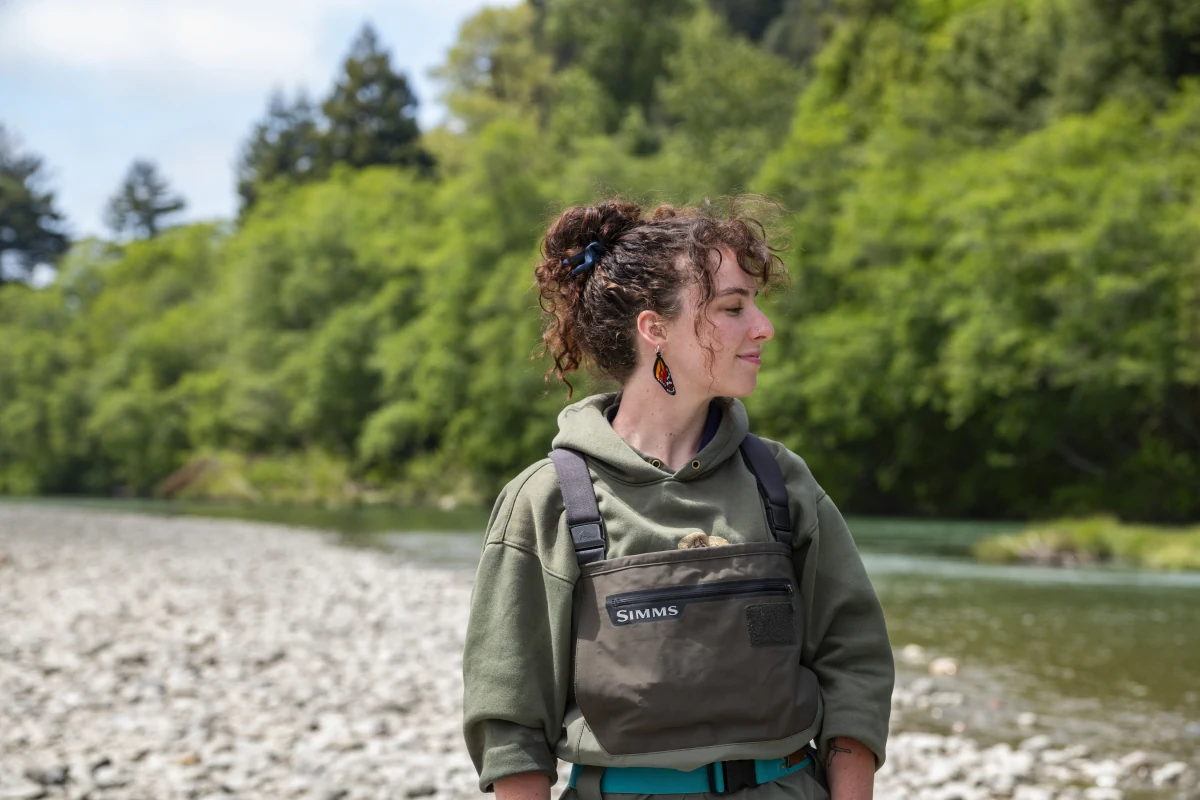 Daphne Cheney stands near a riverbank in waders, looking across the water.