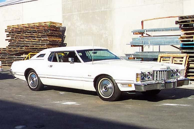 A white 1973 Ford Thunderbird parked beside stacks of lumber.