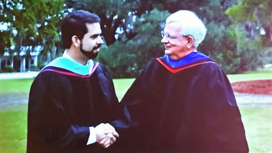 Richard Carvajal and an older man in academic regalia shake hands outdoors.