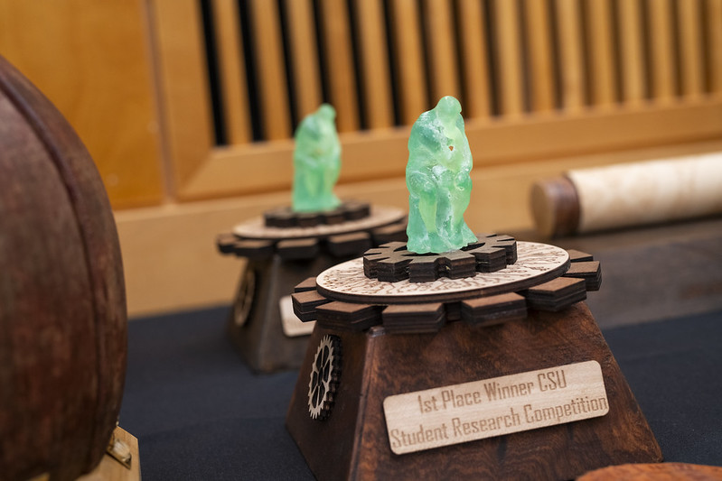 A photo of the 1st Place Winner trophy for the 39th Annual CSU Research Competition. This trophy features a sea foam green thinking man figure mounted to a wooden base with gear-like detailing. A plaque on the front reads "1st Place Winner CSU Student Research Competition." Another identical trophy is visible in the background.