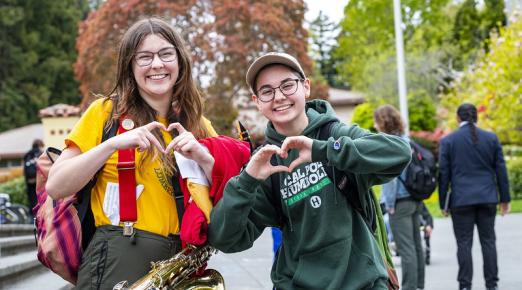 A photo of two students holding up their hands as hearts on campus.