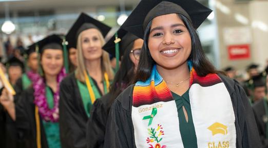 A female graduate smiling, dressed in a black cap and gown with a Hispanic stole that reads "Gonzalez." 