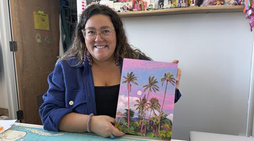 Gina Tuzzi at a desk, holding up her painting of palm trees against a pink and purple sky.