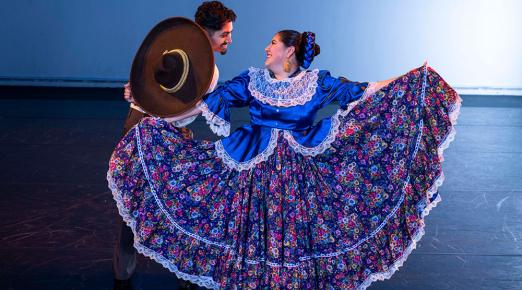 male and female folklorico dancers looking at teach other, male is holding up a hat and female is spreading her colorful floral skirt. 