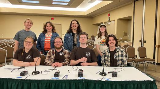 4 members of the quiz bowl team sit at a table ready to compete, with 4 supporting members standing directly behind them. 