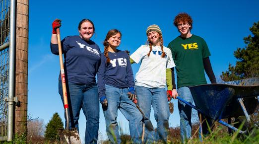 four students stand side-by-side smiling and holding shovels, wearing clothing with the Y.E.S. program logo on it.