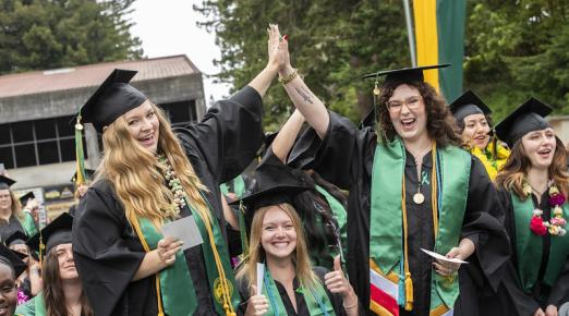 A photo of three graduates at commencement with two of them high fiving each other above the third graduate sitting down. .