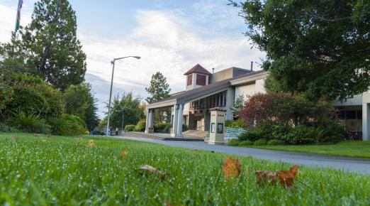 A view of the front of the Cal Poly Humboldt Library with dewy green foliage at the forfront and trees and a blue sky framing the shot.