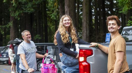 A student sits in the back of a flatbed truck full of her belongings. Two others are with the student - one is wearing a t-shirt that says "Cal Poly Humboldt Dad."