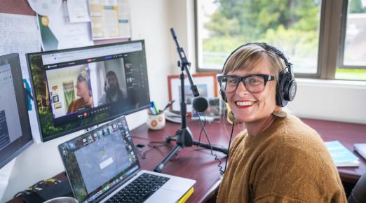 Sarah Ray sitting at desk with a computer screen in the background and a mic near her face