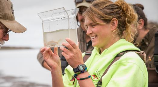 a student, blonde hair tied back in a pony tail, smiles while holding a a water sample with a fish in it.