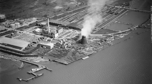 black and white aerial shot of an energy plant along the water, smoke bellowing out from a tower.