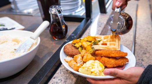 A photo of someone pouring maple syrup on a full plate of waffles, chicken, eggs during Late Night Breakfast.