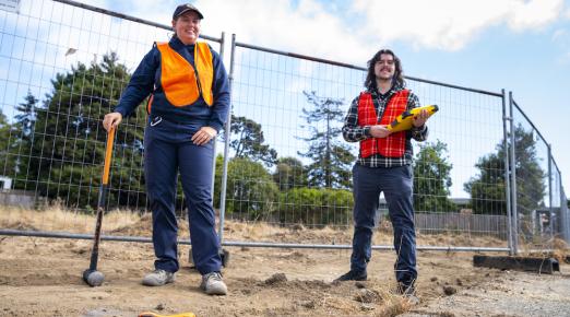 two students standing conducting researching, one holding a sledgehammer and the other, a clipboard.