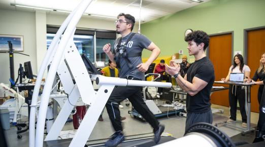 A photo of a student in the Kinesiology Human Performance Lab running on a treadmill wearing a vest that tracks their movement while another student cheers them on. 