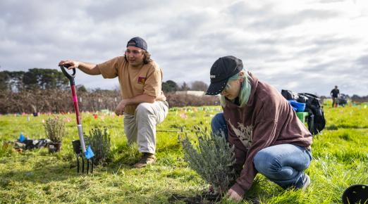 One student holding a shovel and another placing a plant into the ground at the campus farm