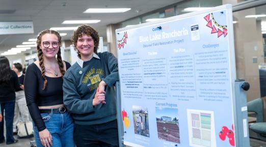 A photo of two students standing in front of their poster presentation in the Library during ideaFest in 2025. 