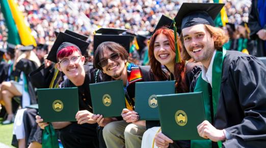A photo of four graduates sitting down holding up their diploma covers on Commencement.