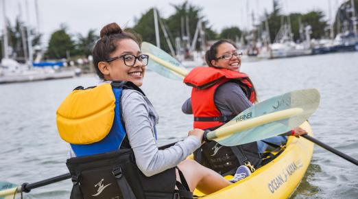 2 people paddling a kayak, looking over their shoulders and smiling. 