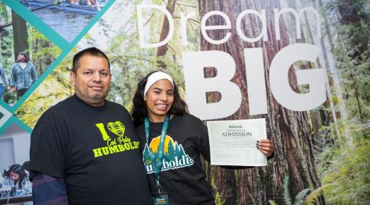 Parent and daughter standing in front of sign reading "Dream Big" holding an admittance letter. 