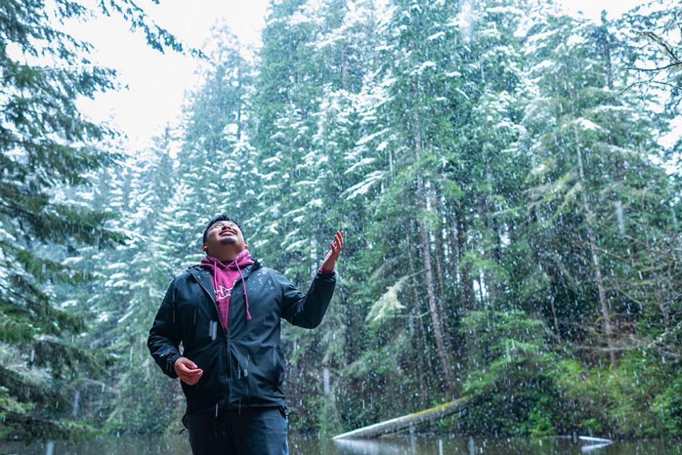A student watches snow fall in the forest behind campus