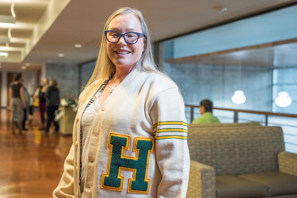 Robin Meiggs smiles at camera while wearing a letterman sweater adorned with a green and gold H