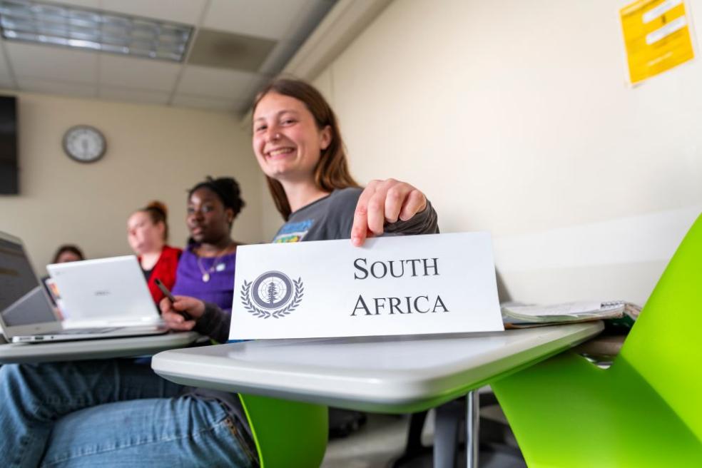 A photo of a student holding a sign with South Africa written on it to represent the country in Model UN. 