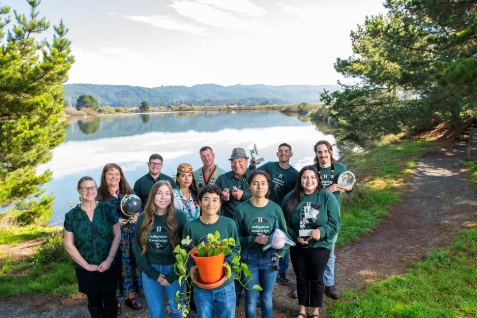 A photo of INRSEP staff gathered at the Arcata Marsh holding a plant, globe, microscope, abalone shell, and plush shark, and wearing matching shirts that state "Indigenize knowledge."