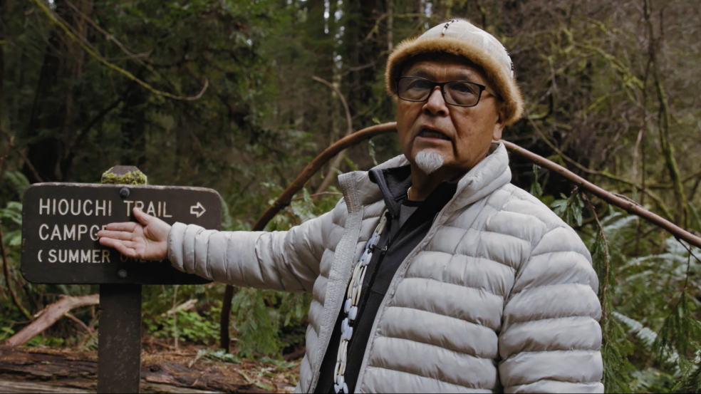 screen from film, Loren Me'-lash-ne Bommelyn stands in front of sign in forest area