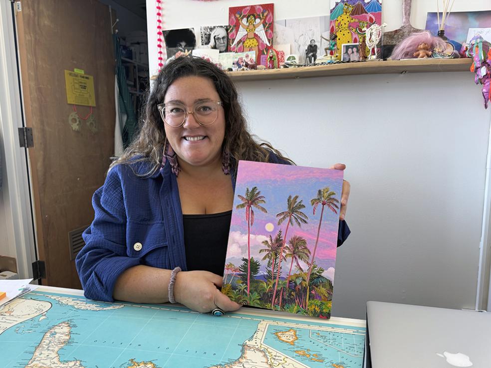 Gina Tuzzi at a desk, holding up her painting of palm trees against a pink and purple sky.