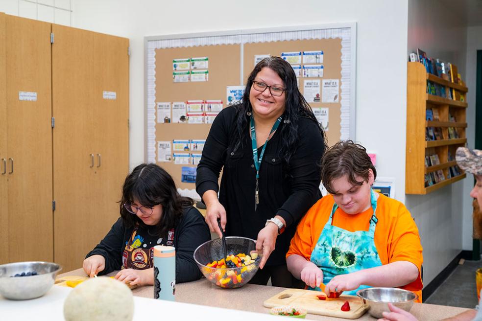 Resident teacher Valerie Canfield stands between two students holding a bowl of fruit salad and smiling at the camera.