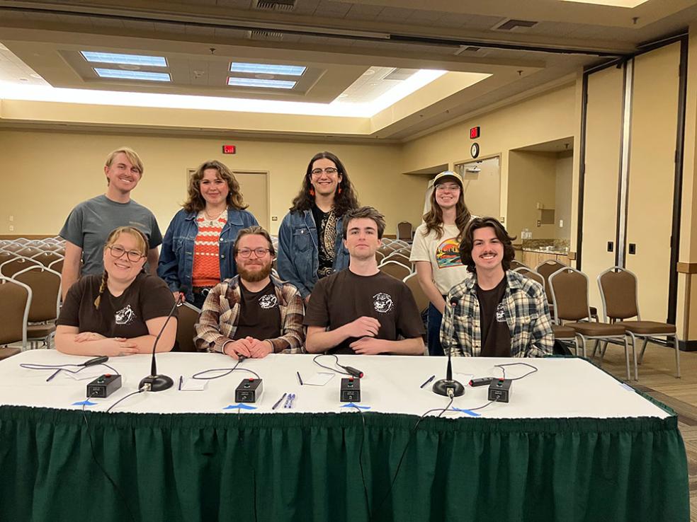 4 members of the quiz bowl team sit at a table ready to compete, with 4 supporting members standing directly behind them. 