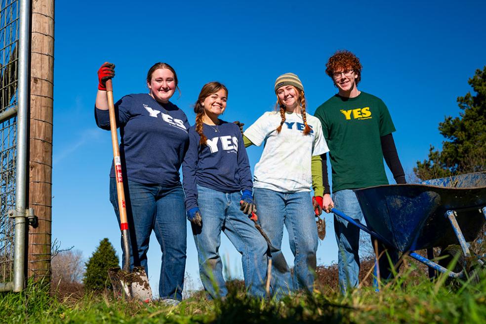 four students stand side-by-side smiling and holding shovels, wearing clothing with the Y.E.S. program logo on it.