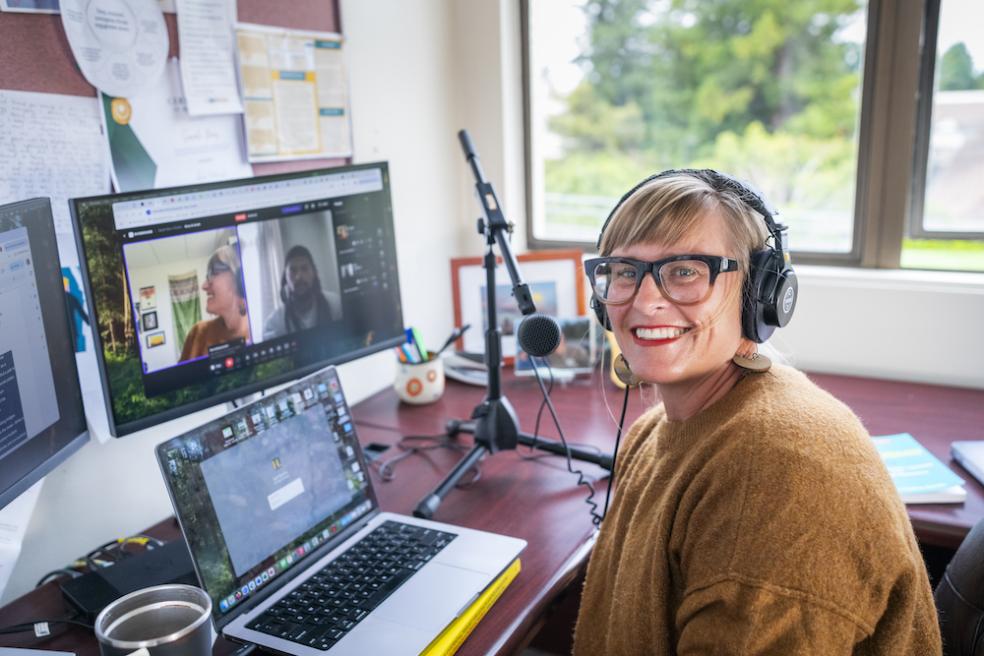 Sarah Ray sitting at desk with a computer screen in the background and a mic near her face