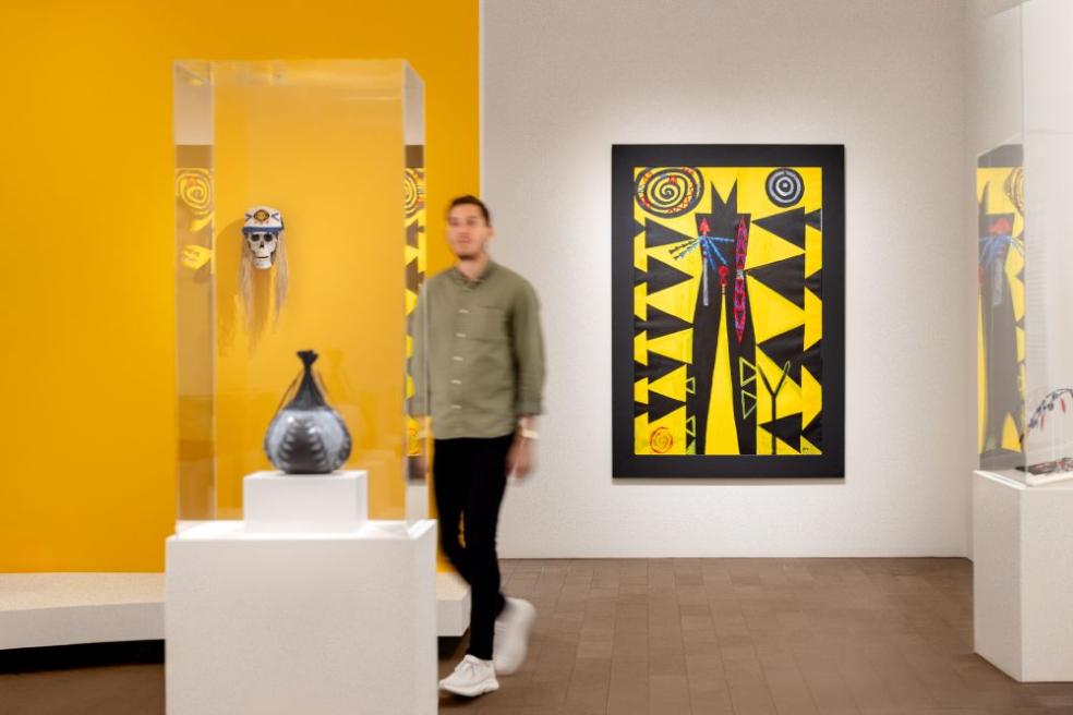 A photo of a blurred person walking through the de Young Museum. The photo features Indigenous artist Brian Tripp's "Someday You Might Have to Fight for What You Believe In" painting in the background.