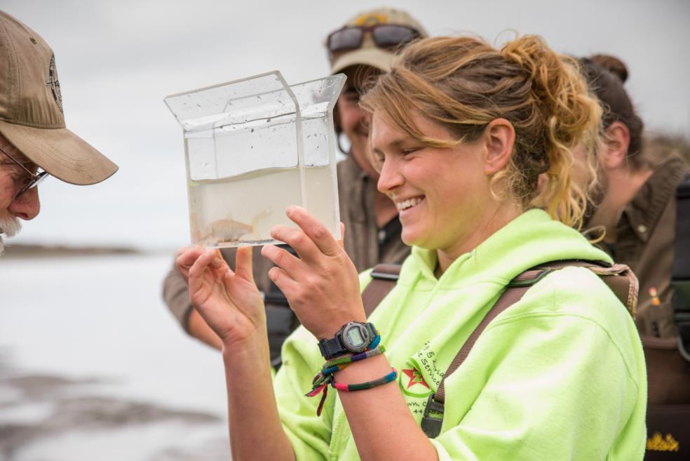 a student, blonde hair tied back in a pony tail, smiles while holding a a water sample with a fish in it. 
