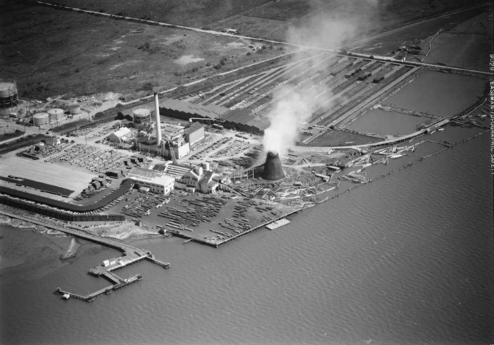 black and white aerial shot of an energy plant along the water, smoke bellowing out from a tower.