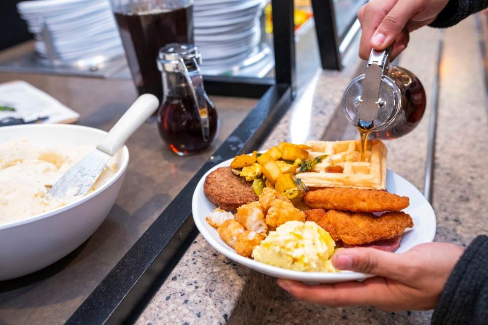 A photo of someone pouring maple syrup on a full plate of waffles, chicken, eggs during Late Night Breakfast.