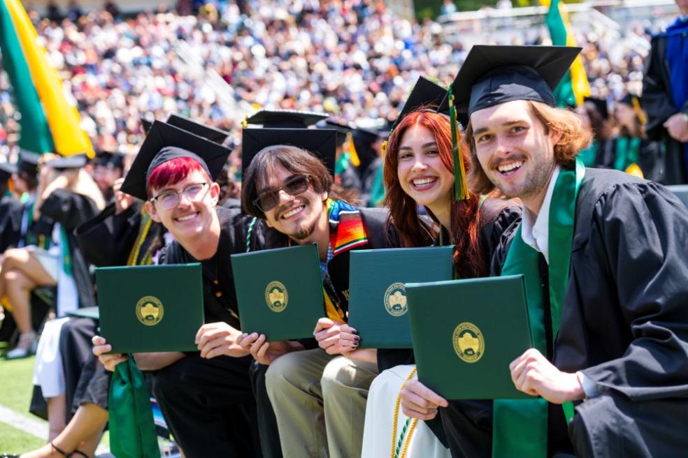 A photo of four graduates sitting down holding up their diploma covers on Commencement.