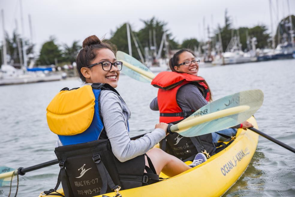 2 people paddling a kayak, looking over their shoulders and smiling. 