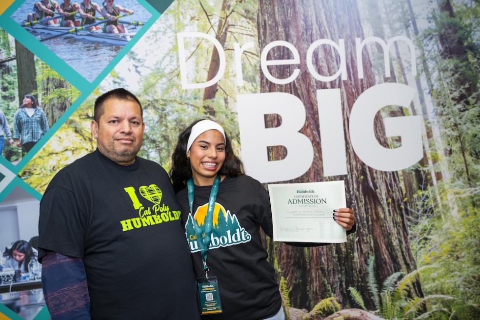 Parent and daughter standing in front of sign reading "Dream Big" holding an admittance letter. 