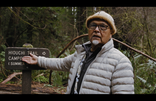 screen from film, Loren Me'-lash-ne Bommelyn stands in front of sign in forest area