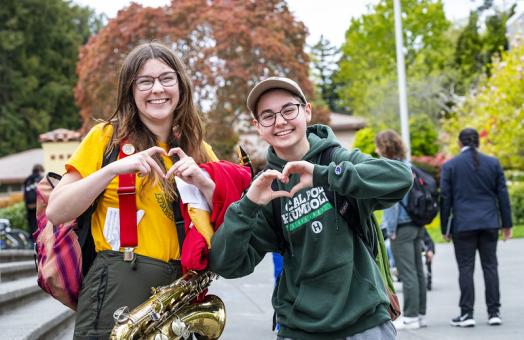 A photo of two students holding up their hands as hearts on campus.