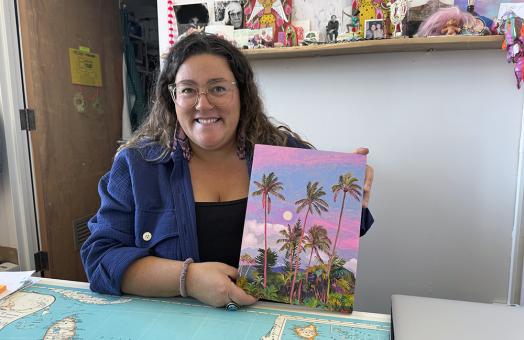 Gina Tuzzi at a desk, holding up her painting of palm trees against a pink and purple sky.